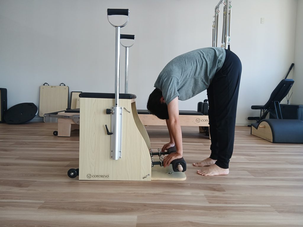ピラティスチェアのバーを両手で押しながら、立位で背骨を丸めて前屈している様子