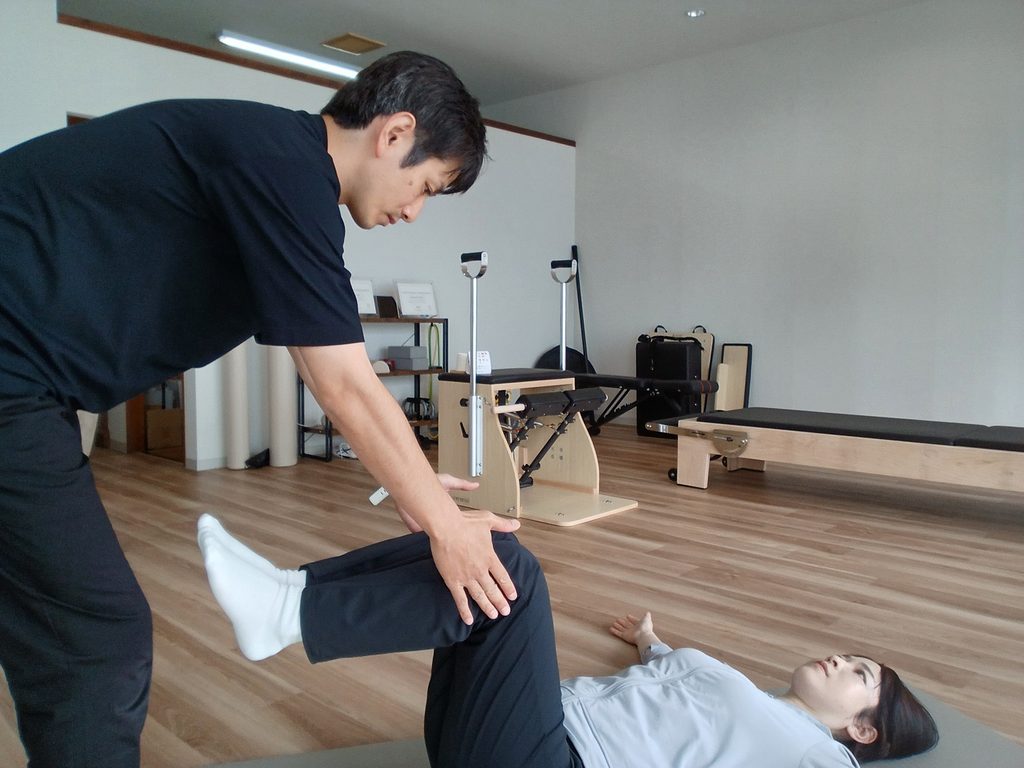 仰向けで両脚を上げ、トレーナーが股関節と骨盤の動きを確認しているAXISのパーソナルレッスン風景。