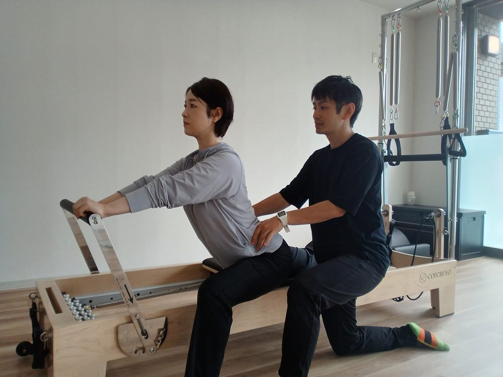 ピラティスマシンのフットバーを持ってランジ姿勢を取り、トレーナーが骨盤に手を添えて姿勢を確認している様子
