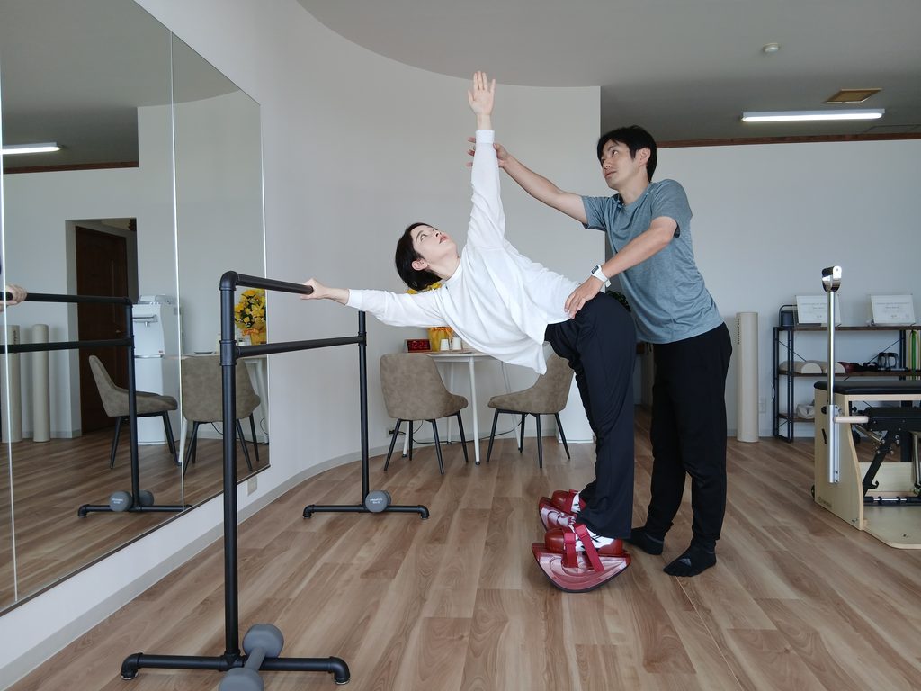 船底状の形をしたシューズを履きながら、バレエバーを持って身体を開くストレッチをしている女性と補助するトレーナー