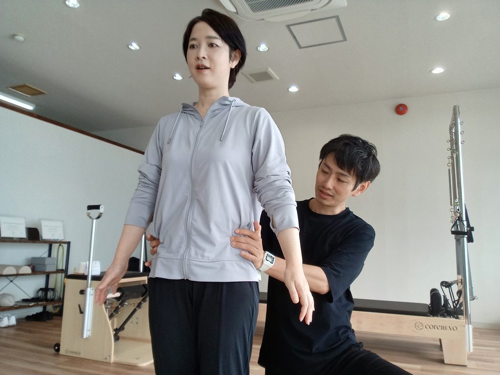 立った姿勢で、トレーナーが体幹と骨盤まわりに手を添え、身体全体の位置関係を確認しているAXISのレッスン風景。
