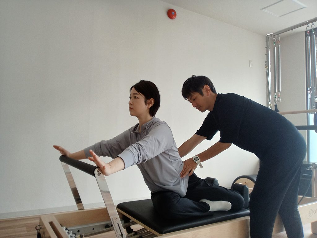 リフォーマーに座って両腕を前へ伸ばし、トレーナーが背中と胸郭まわりに手を添えて身体の位置を確認しているAXISのレッスン風景。