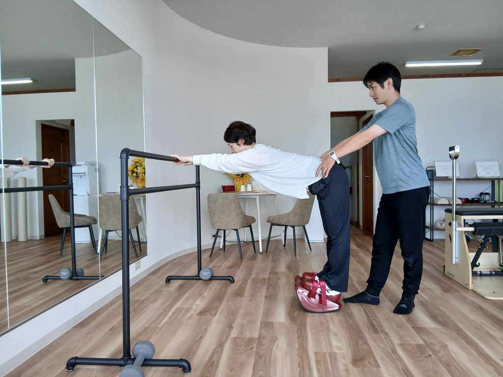 立位で股関節を曲げ、トレーナーが骨盤まわりに手を添えながら身体の動きを確認しているAXISのレッスン風景。
